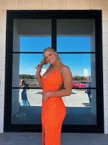 A girl in an orange dress standing in front of a black framed glass door with a reflection of a parking lot and blue sky.