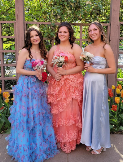 Three young women in colorful ruffle dresses holding bouquets of flowers, standing outdoors in front of a wooden fence with green foliage and tulips.