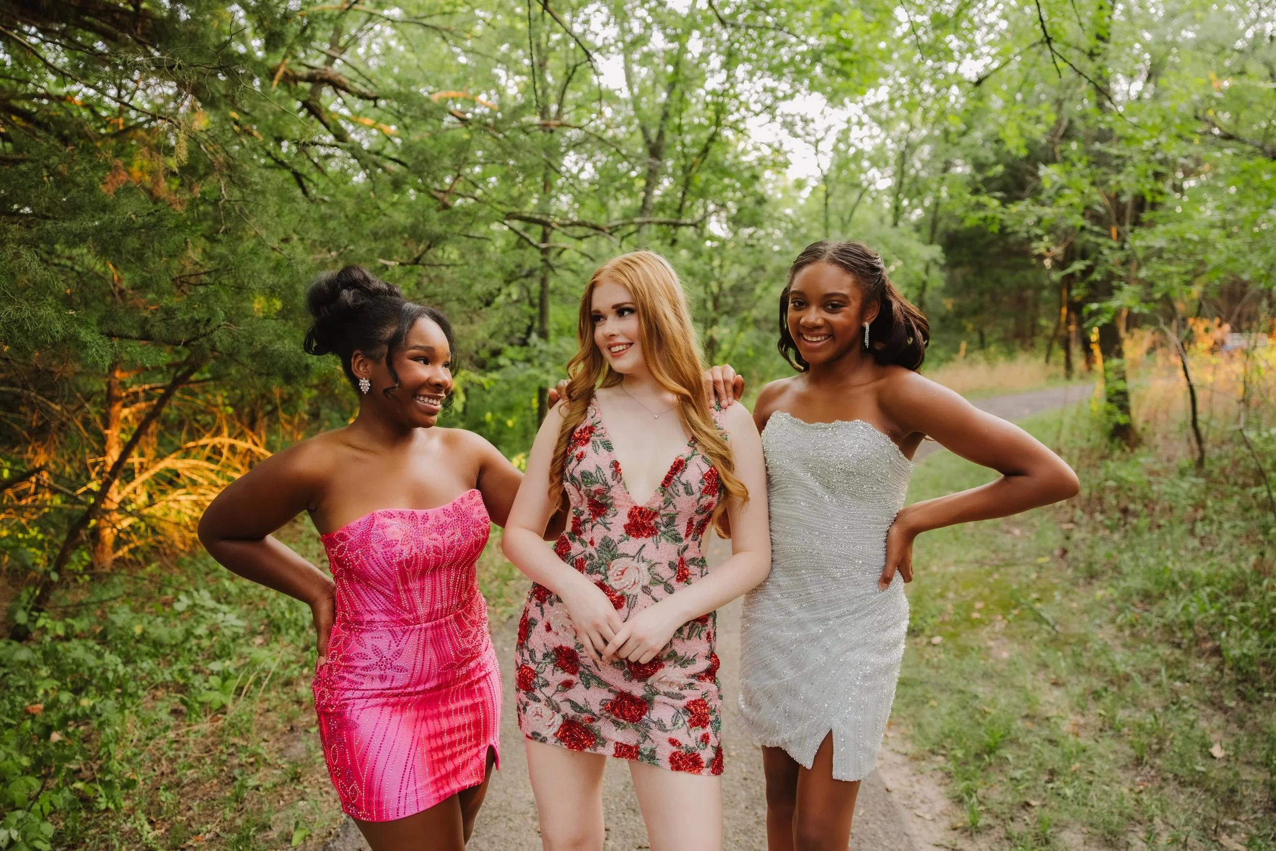 Four young women dressed in colorful formal dresses pose outdoors on grass with trees in the background.