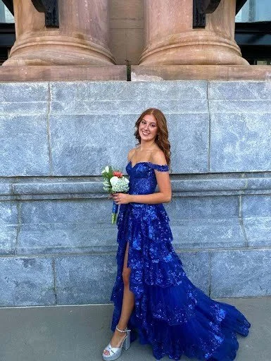 Young woman in a royal blue, off-the-shoulder, strapless Sherri Hill gown holding a bouquet, standing against a stone wall with classical columns in the background.