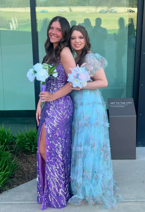 Two young women in colorful dresses holding bouquets of flowers, smiling and posing outside near a glass building.