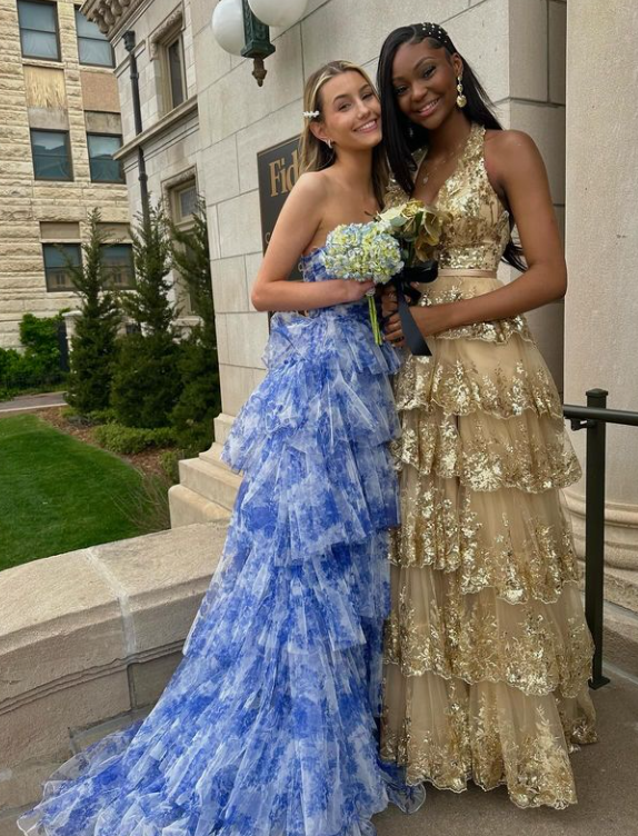 Two young women in formal dresses standing on stairs outside a building, smiling and hugging. One in a blue ruffled gown holding a bouquet, the other in a gold ruffled gown.