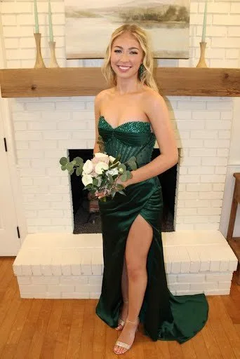 A young woman in a green strapless dress with a high slit, holding a bouquet of flowers, standing in front of a fireplace