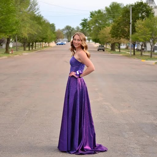 A young woman in a purple, strapless, floor-length gown standing on a quiet street with trees and cars in the background.