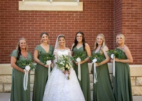 Group of six women, including a bride in a white wedding dress holding a bouquet, and five bridesmaids in green dresses holding bouquets, standing outside in front of a brick wall.