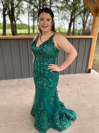 Young woman in a teal lace evening gown posing outdoors on a wooden deck, with trees and a fence in the background.