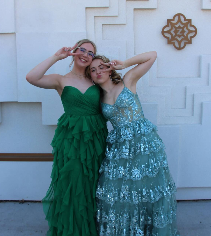 Two young women in formal dresses posing for a photo in front of a white decorative wall. One is wearing a strapless green dress with ruffles, and the other is wearing a blue lace dress with floral patterns. They are making peace signs with their han