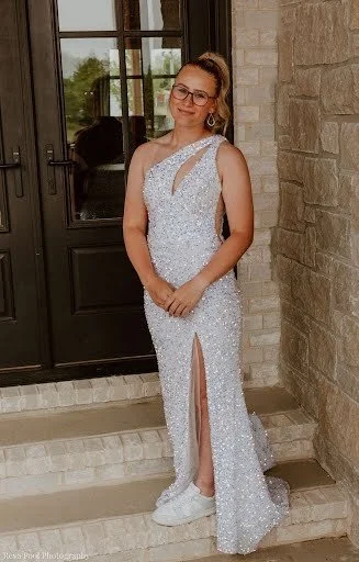 Young woman in a sparkly, silver gown with a high slit, sitting on steps outside a building with a stone wall and glass door, smiling at the camera.