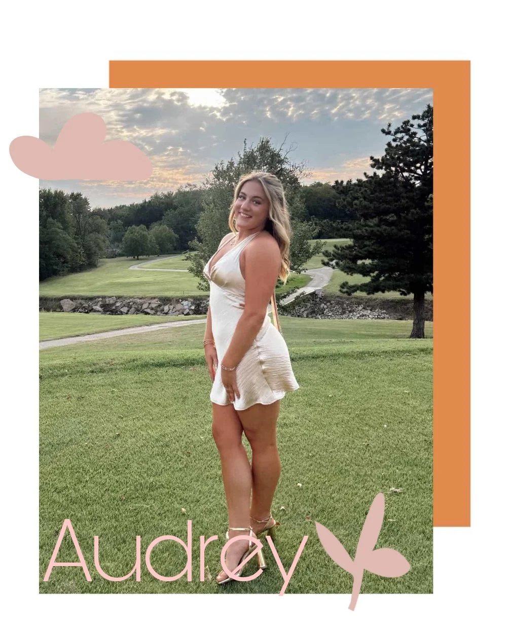 A young woman in a cream-colored dress standing on a grassy field with trees and a winding path in the background, under a partly cloudy sky during sunset, with the name 'Audrey' and decorative pink leaves and a pink cloud overlay.