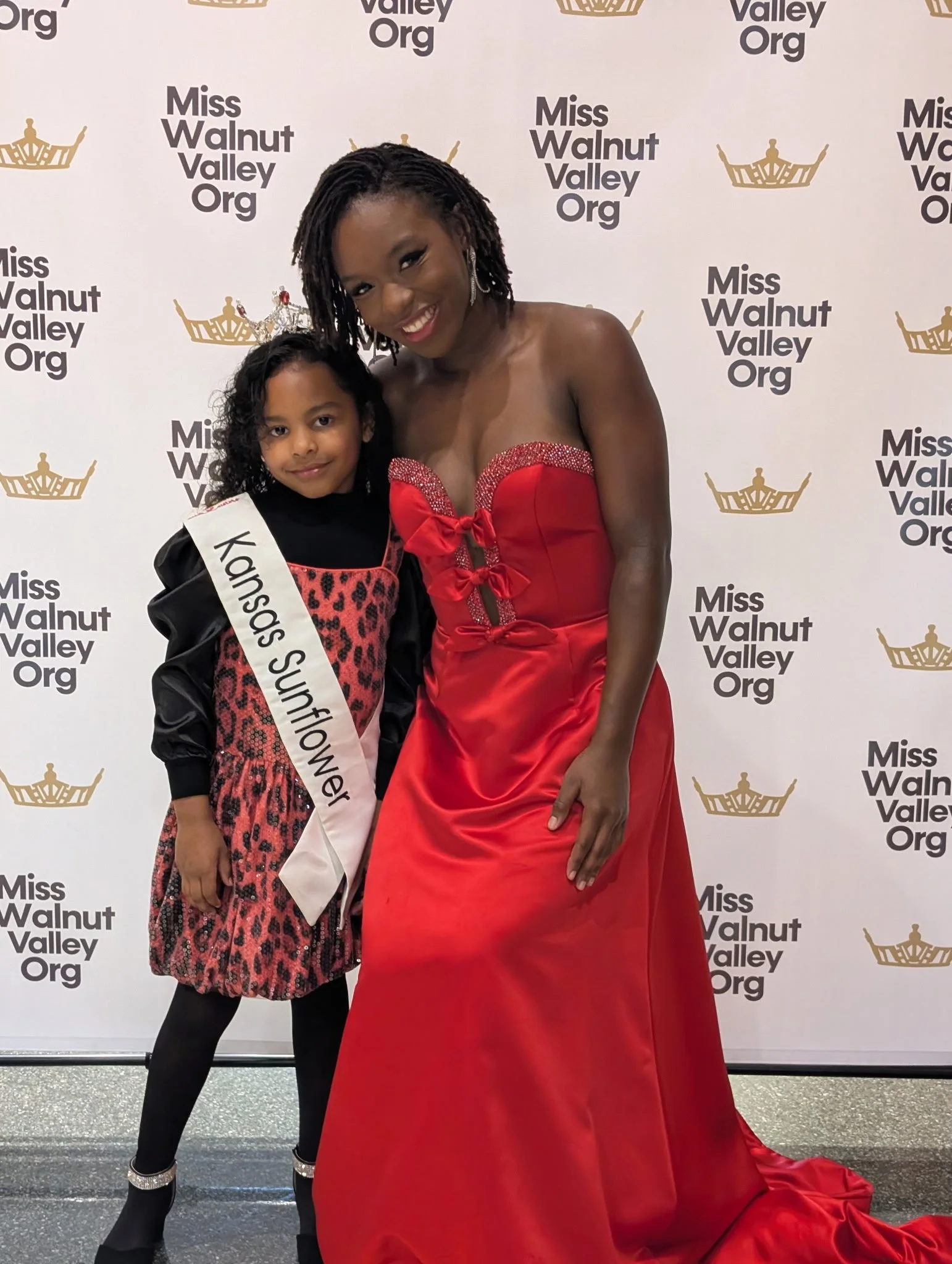 A young girl wearing a sash and a black and red leopard print dress stands next to a woman in a strapless red gown at a Miss Walnut Valley Organization event.