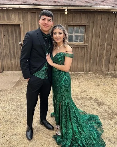 A young man and woman dressed in formal attire, standing outside in front of a wooden fence, with the woman wearing a long green gown and the man in a black suit.