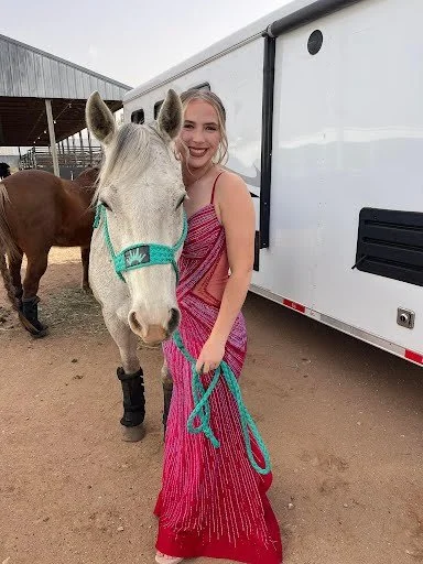 A woman in a pink dress standing next to a white horse with a green halter at a stable.