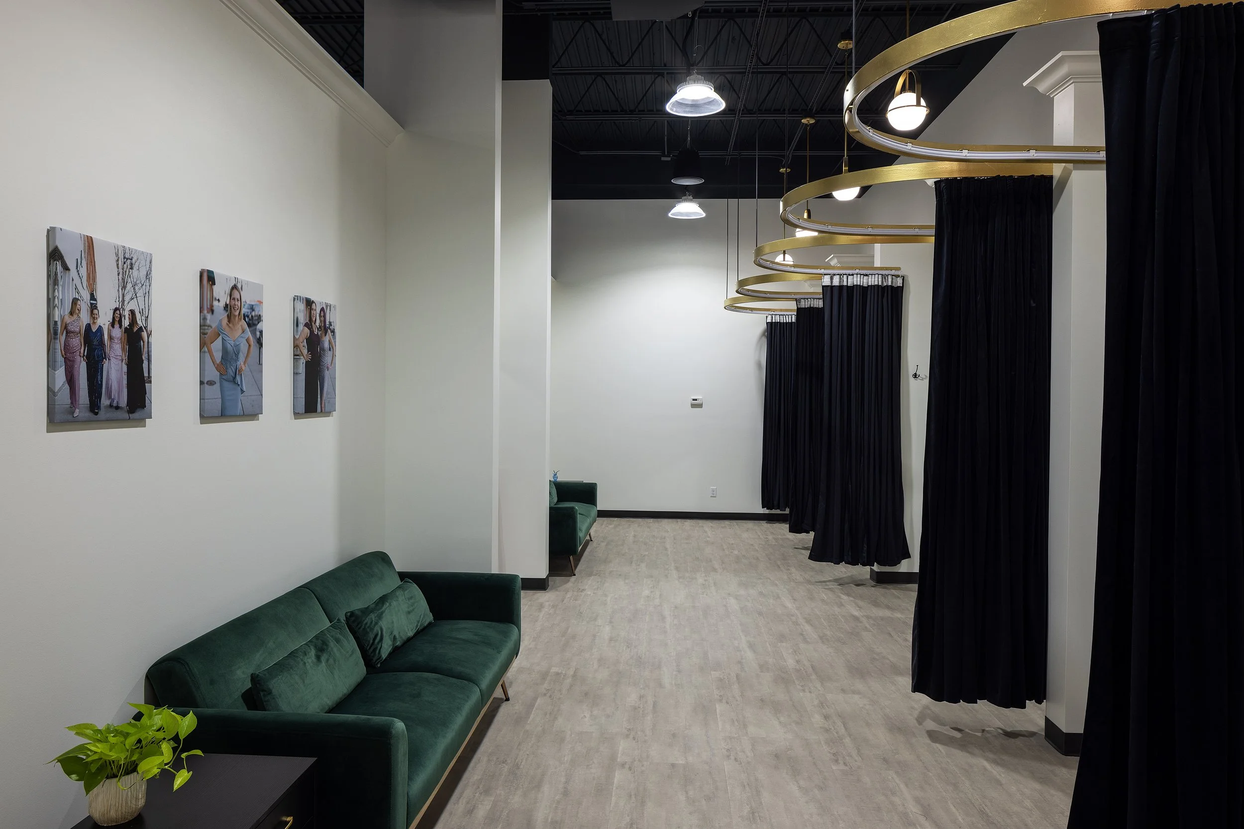 An interior view of a fitting room area with black curtains, a green sofa, a small table with a plant, and framed photos on a white wall.