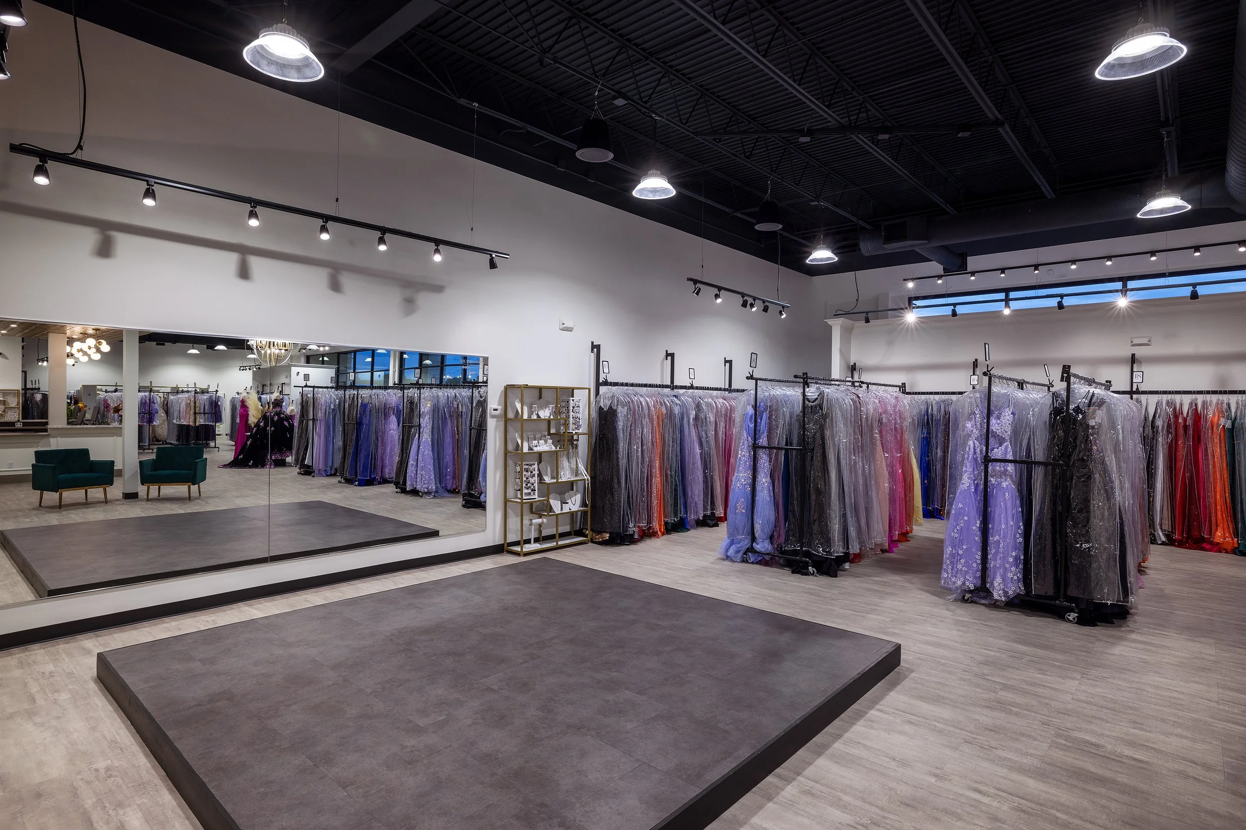 Fitting room with racks of colorful evening gowns covered in plastic, large mirror, and seating area.