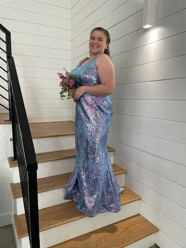A young woman in a shiny, purple-blue sequined gown holding a bouquet of flowers, standing on a staircase with white shiplap walls.