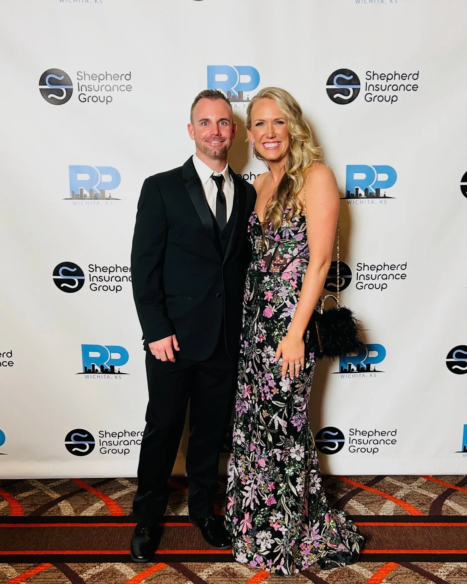 A man and woman dressed formally, standing in front of a step and repeat banner with logos for Shepherd Insurance Group and RP Wichita, KS, at an event.