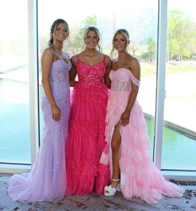 Three young women in elegant pink and purple evening gowns posing in front of a large window overlooking a lake and trees.