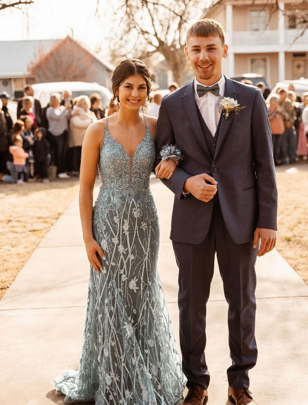 Young couple in formal attire standing on a sidewalk at an outdoor event, with people in the background.