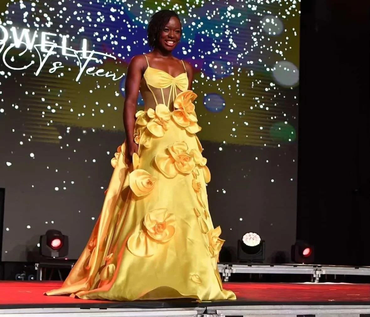 A woman on stage wearing a yellow gown with large flower decorations, smiling with a dark background and stage lights.