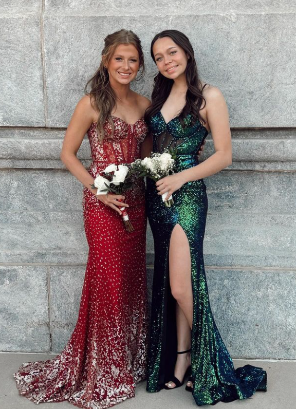 Two young women in formal gowns standing against a stone wall, holding white flower bouquets and smiling.