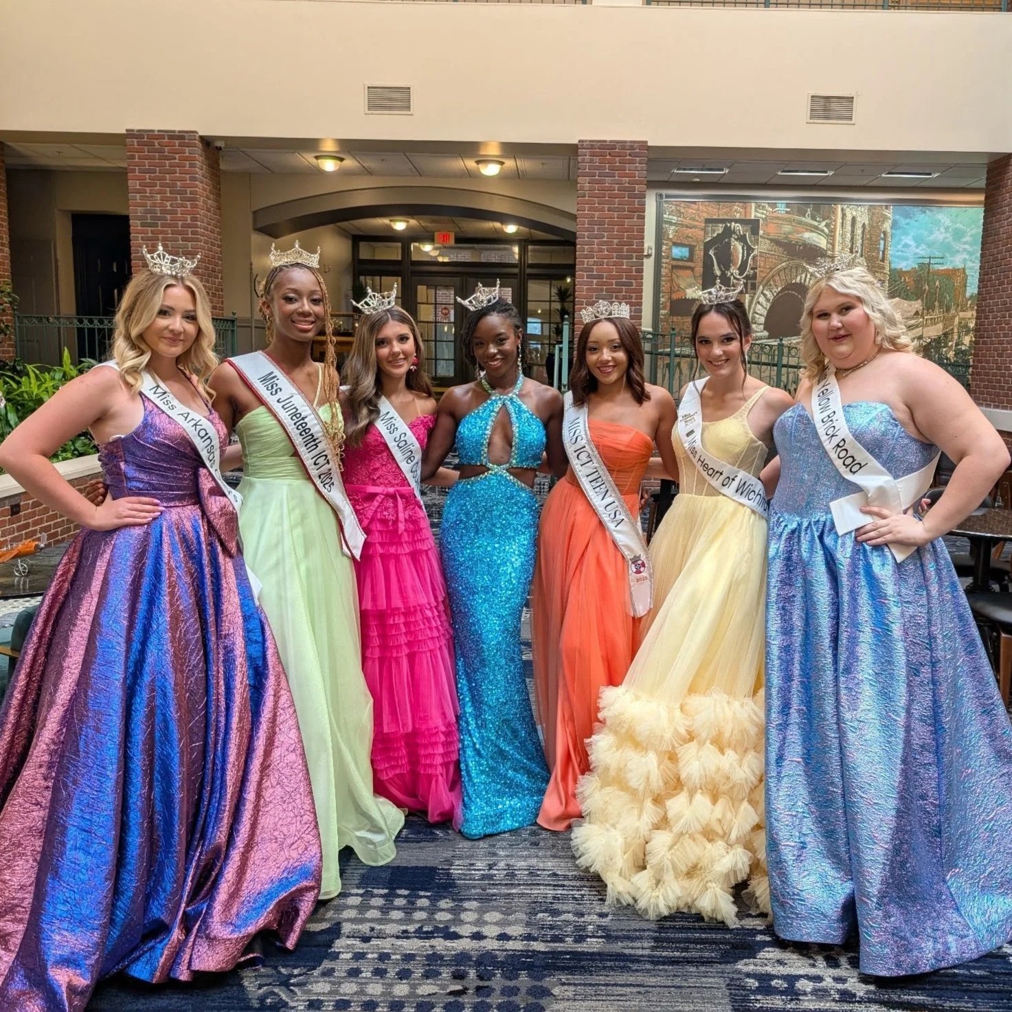 Seven young women wearing colorful evening gowns and tiaras, posing together indoors, each with a sash indicating they are pageant winners.