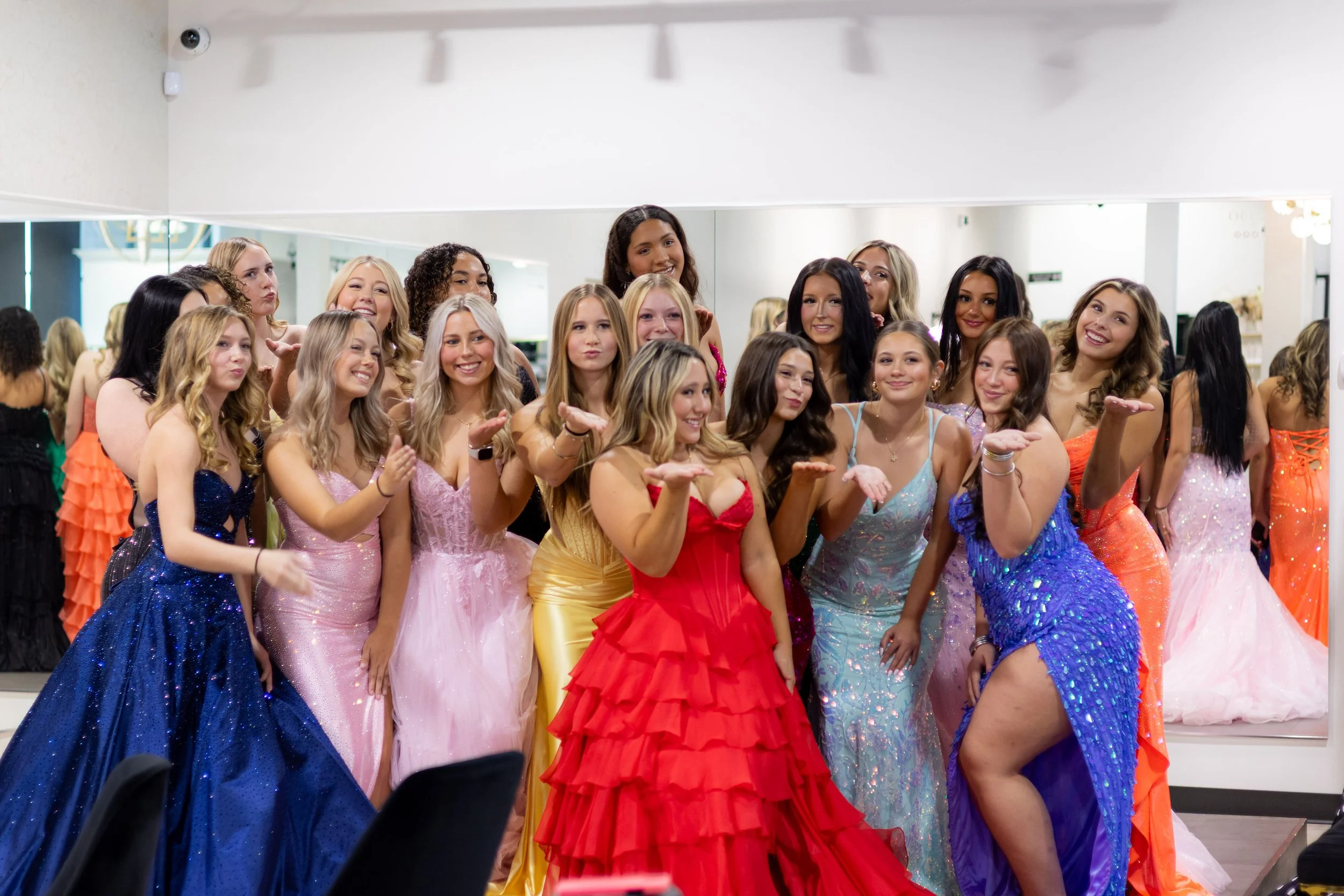 A group of young women in colorful formal dresses posing together in front of a mirror in a dressing room.