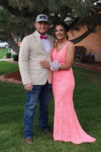 A young man and woman dressed in formal attire standing outdoors in front of a tree, smiling at the camera.