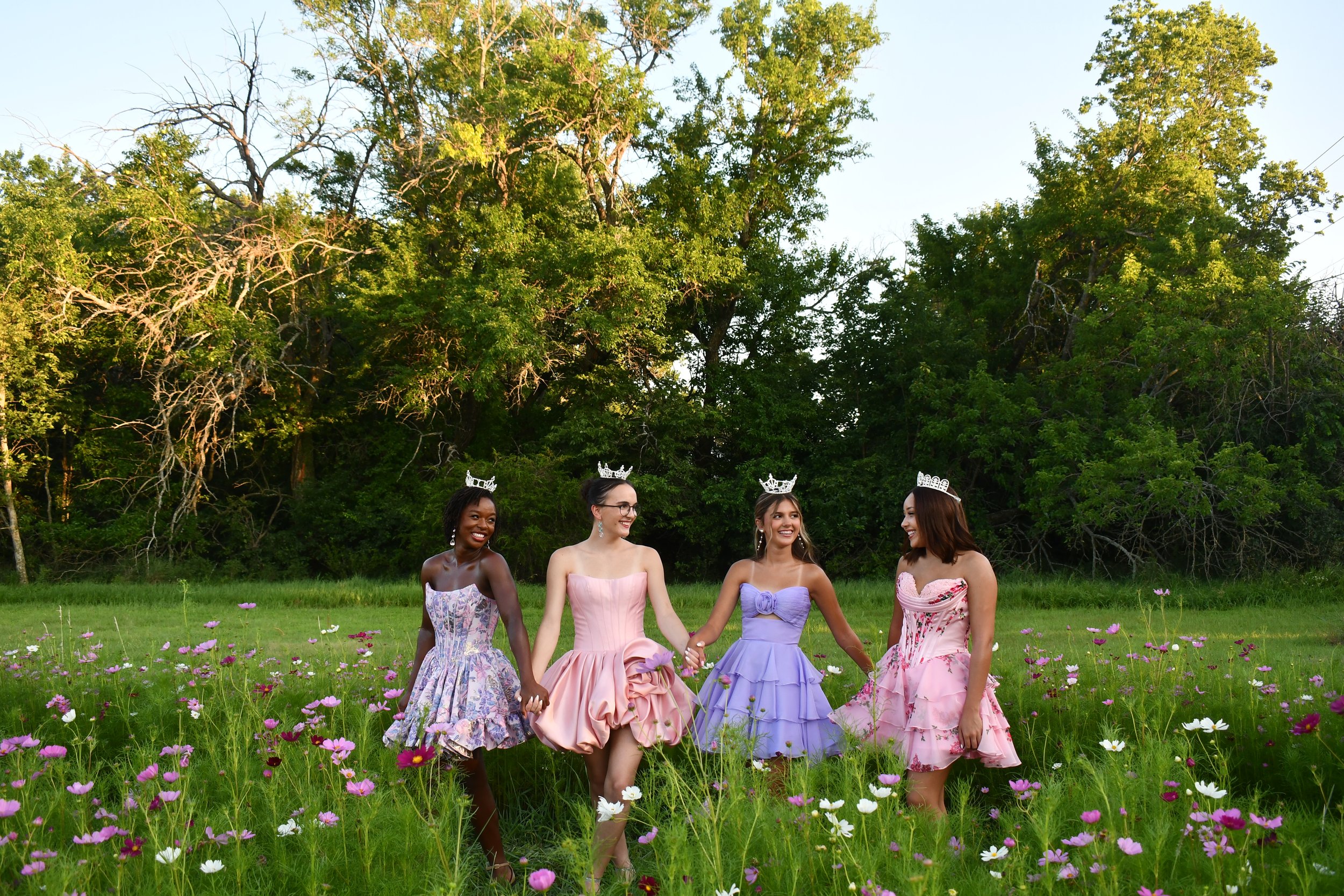 Four young women dressed in colorful formal dresses pose outdoors on grass with trees in the background.