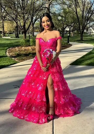 Young woman in a pink off-shoulder ball gown with floral lace, standing on a sidewalk in a park, holding a bouquet of flowers, smiling.
