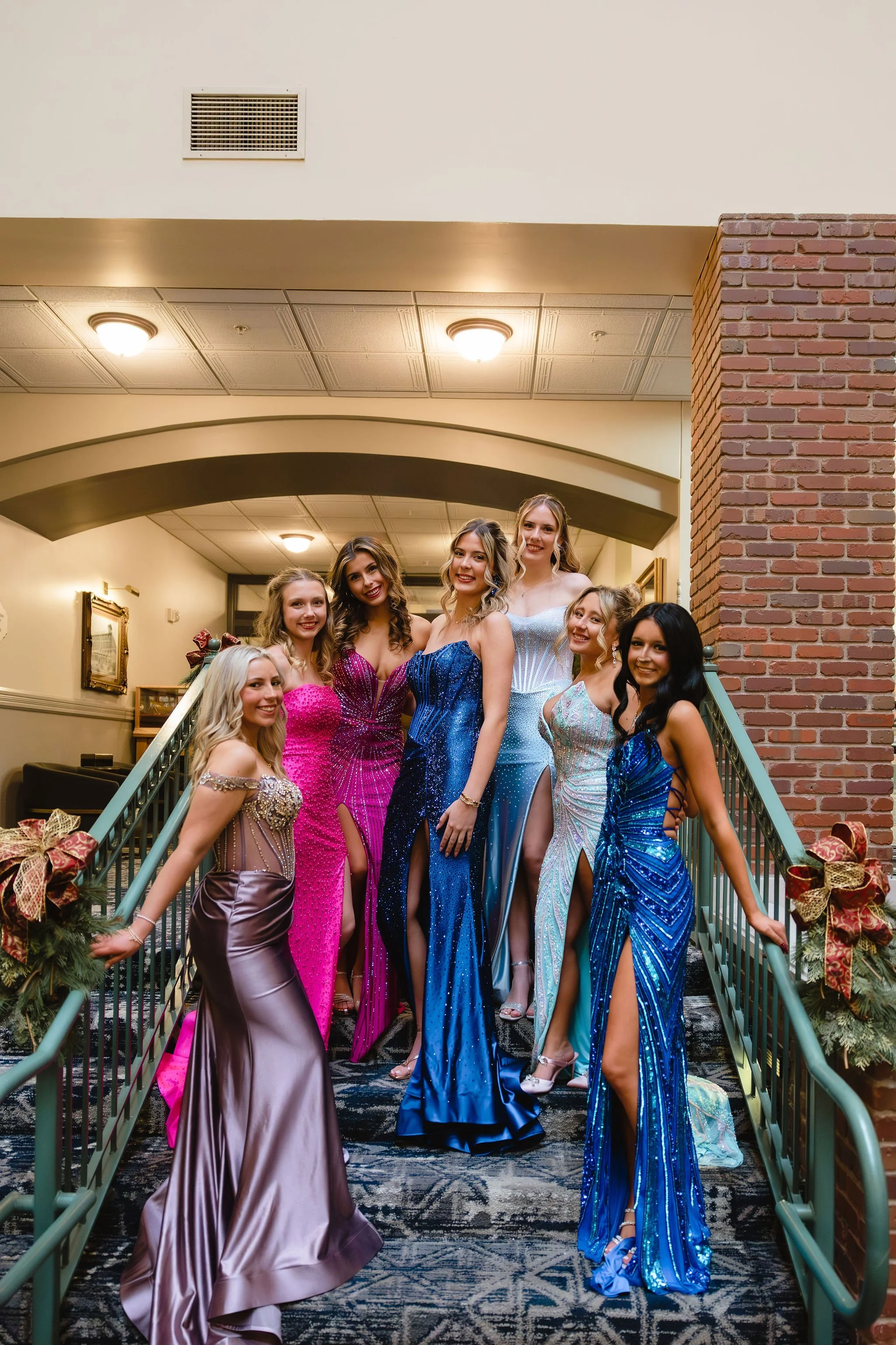 Group of young women in colorful prom dresses posing on a staircase inside a historic building with ornate architecture.