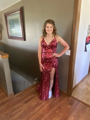 A young woman in a pink and red sequined gown standing in a hallway at home, smiling and posing with one hand on her hip.