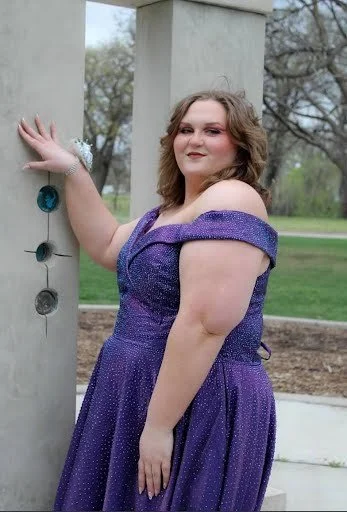 A young woman in a purple dress standing outdoors near a concrete pillar with circular holes.