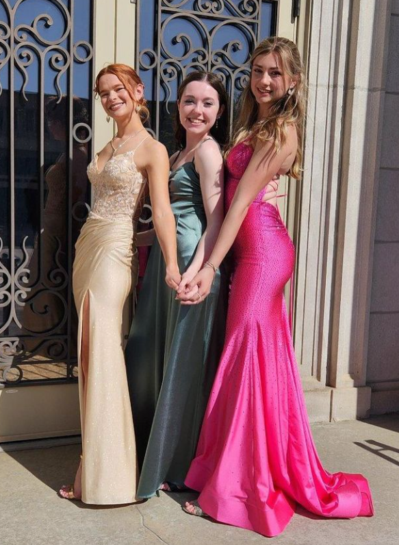 Three young women in formal evening gowns standing outside, holding hands, and smiling at the camera.