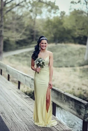 A woman in a yellow gown holding a bouquet, standing on a wooden bridge with a natural landscape background.