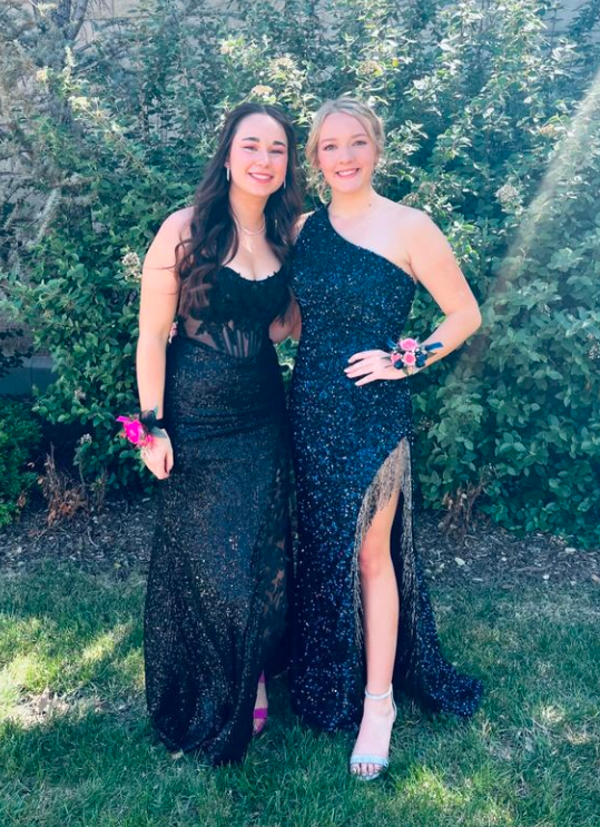 Two young women in formal evening dresses standing outdoors on grass, smiling, with bushes and trees in the background.