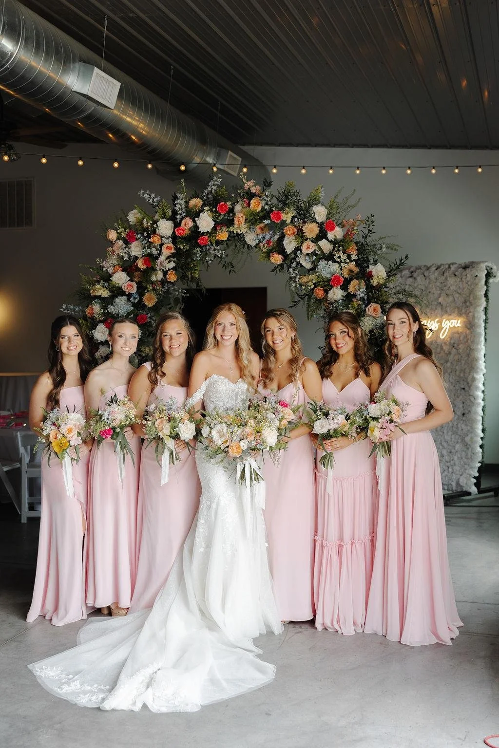 Bride and five bridesmaids standing outside in front of a brick wall, holding bouquets and dressed in green dresses.