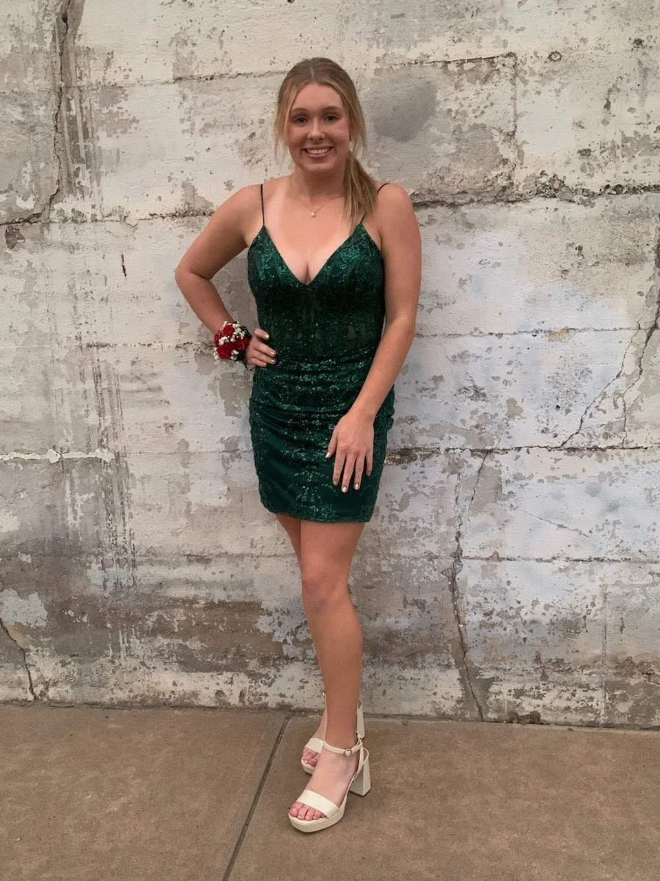 A young woman in a green sequined dress with spaghetti straps, standing against a textured, weathered concrete wall, smiling with one hand on her hip, wearing beige high heels.