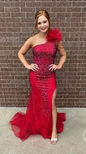 Young woman in a red, sequined gown with a high slit and a large red flower on her shoulder, standing against a brick wall.