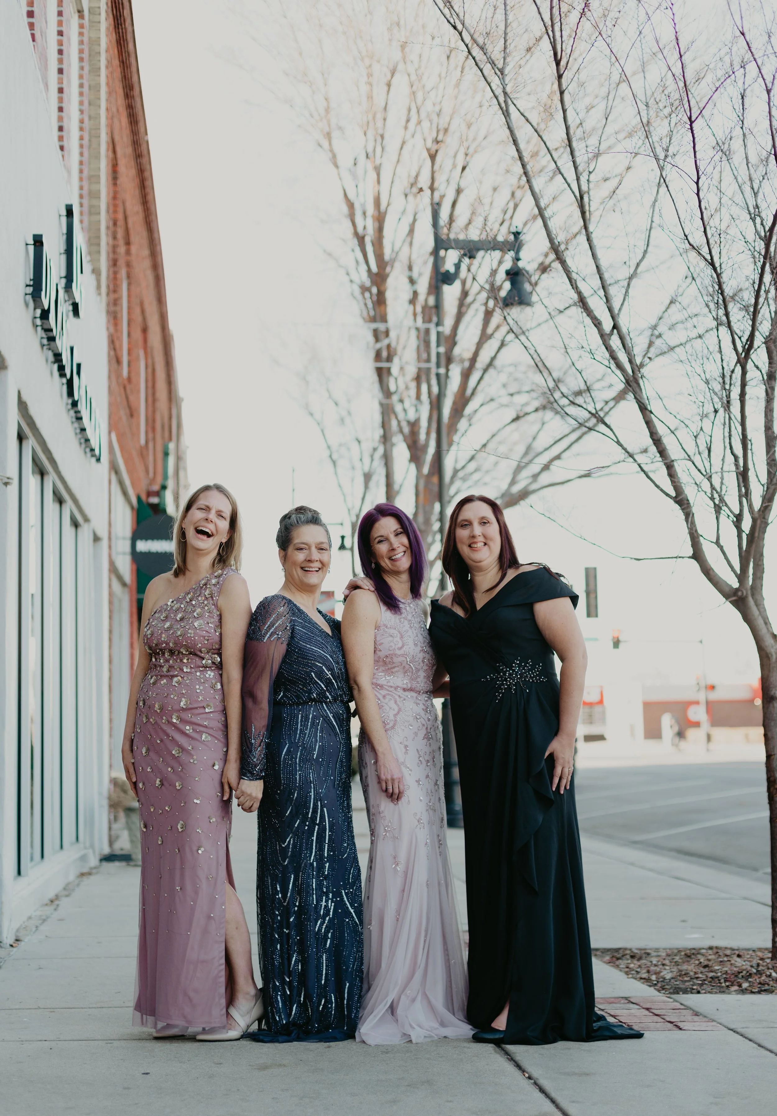 Four women in formal dresses standing on a city sidewalk during daytime, smiling and posing for a photo.