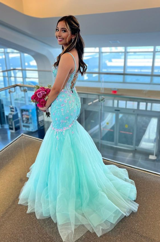 A woman in a light blue, elegant mermaid-style gown with intricate embellishments, holding a bouquet of pink roses, inside a modern building with large windows and a view outside.