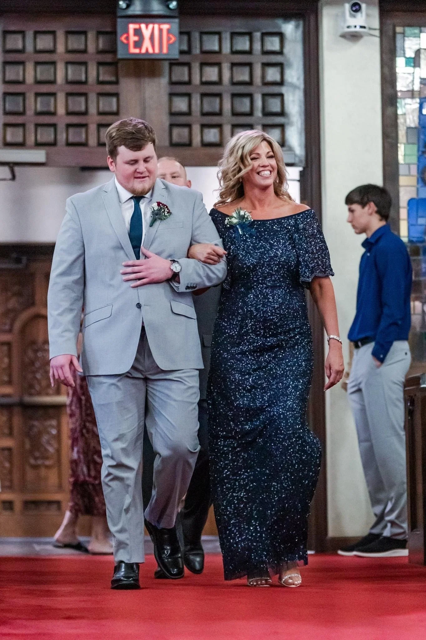 A woman in a glittery navy blue mother of the occasion dress walking down the aisle with a man in a light gray suit. They are inside a church with stained glass windows, wood paneling, and an illuminated exit sign above.