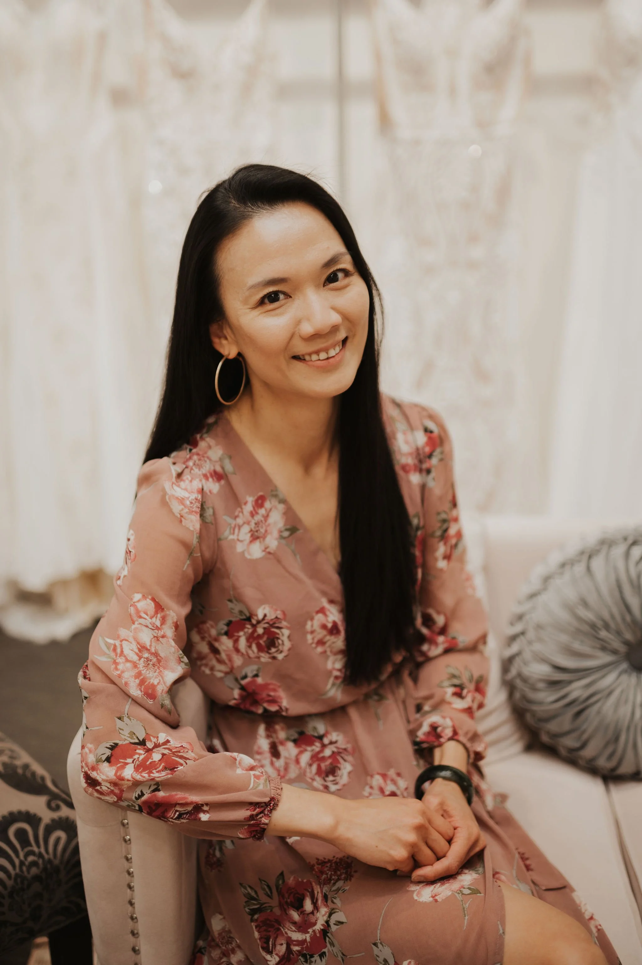 A woman with long black hair, wearing hoop earrings and a floral pink dress, sitting on a couch indoors and smiling at the camera.