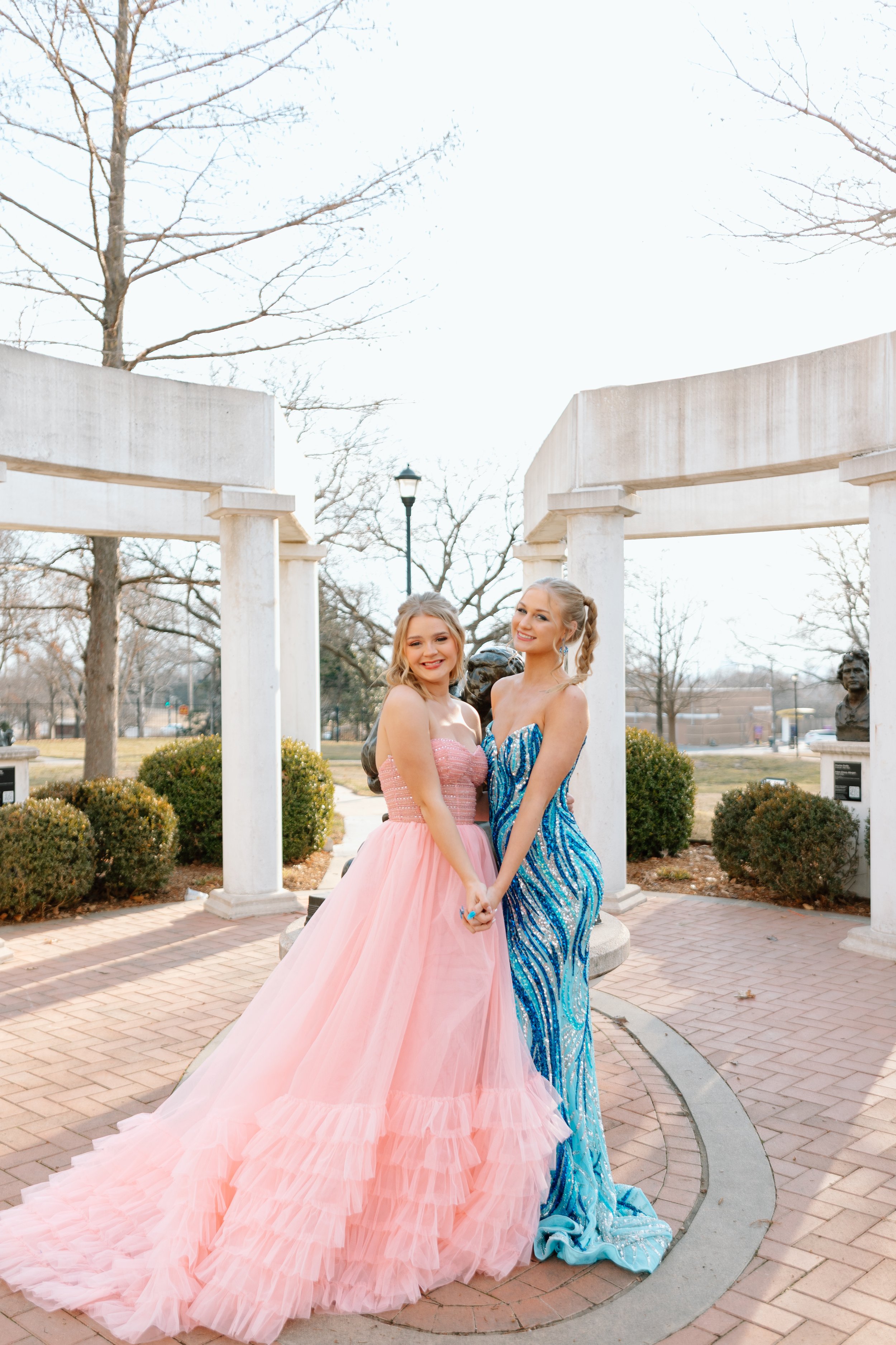 Two young women in formal dresses standing together outdoors, holding hands. One is in a pink ball gown with a ruffled skirt, and the other in a blue and silver sequined evening gown. They are smiling and posing in front of a decorative white structure with columns, trees, and a lamppost in the background.