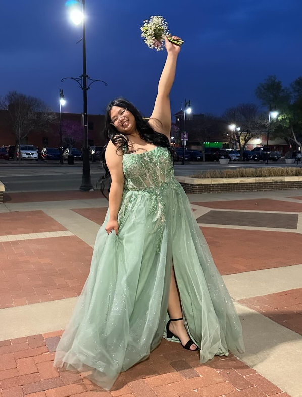 A woman in a long, sparkly, green dress with a slit, standing on a sidewalk at night, holding a bouquet of flowers in the air, smiling, with streetlights and parked cars in the background.