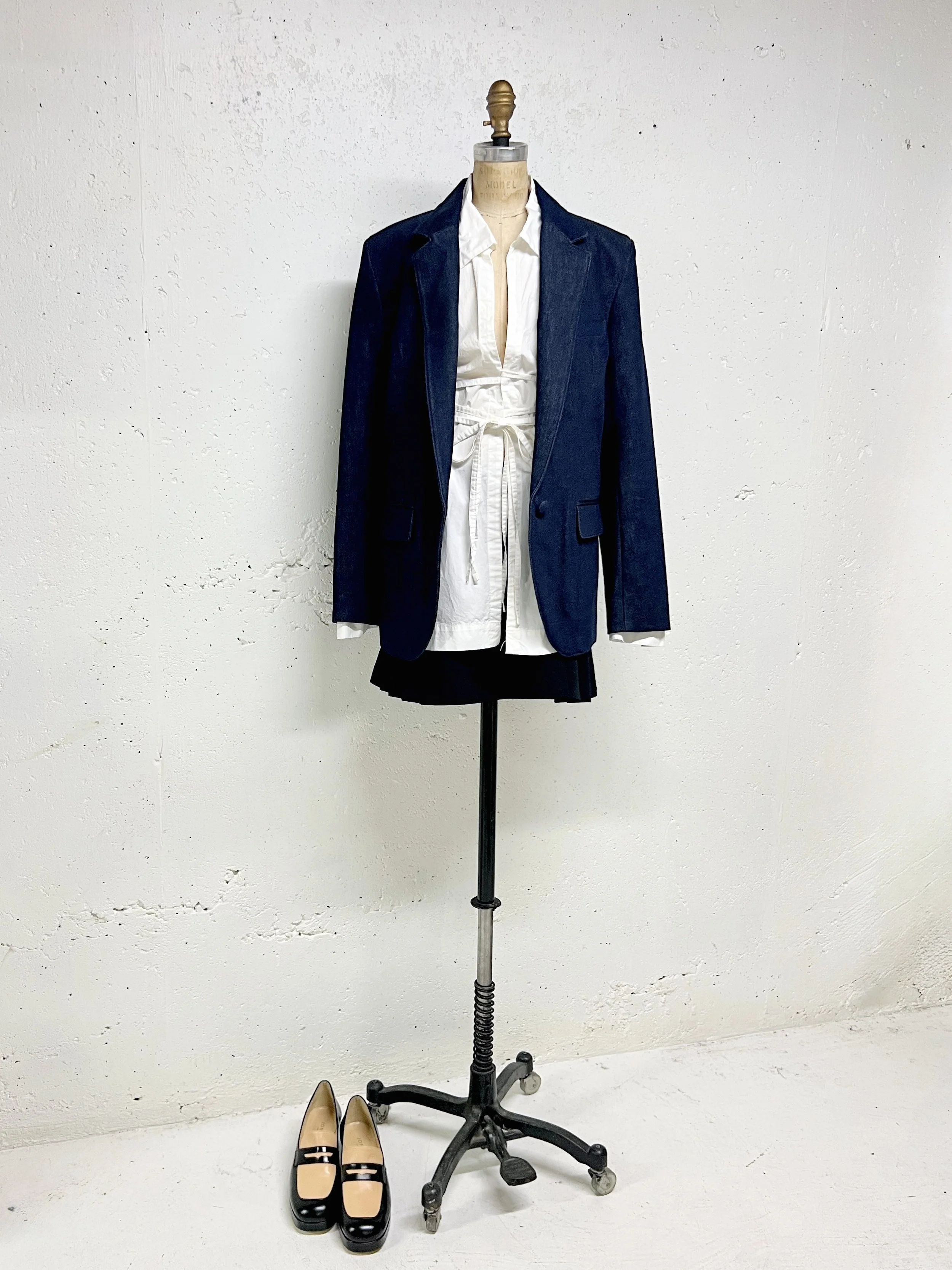 Mannequin dressed in a white shirt, black skirt, and navy blazer, with a pair of black and beige shoes placed on the floor beside it against a plain white wall.