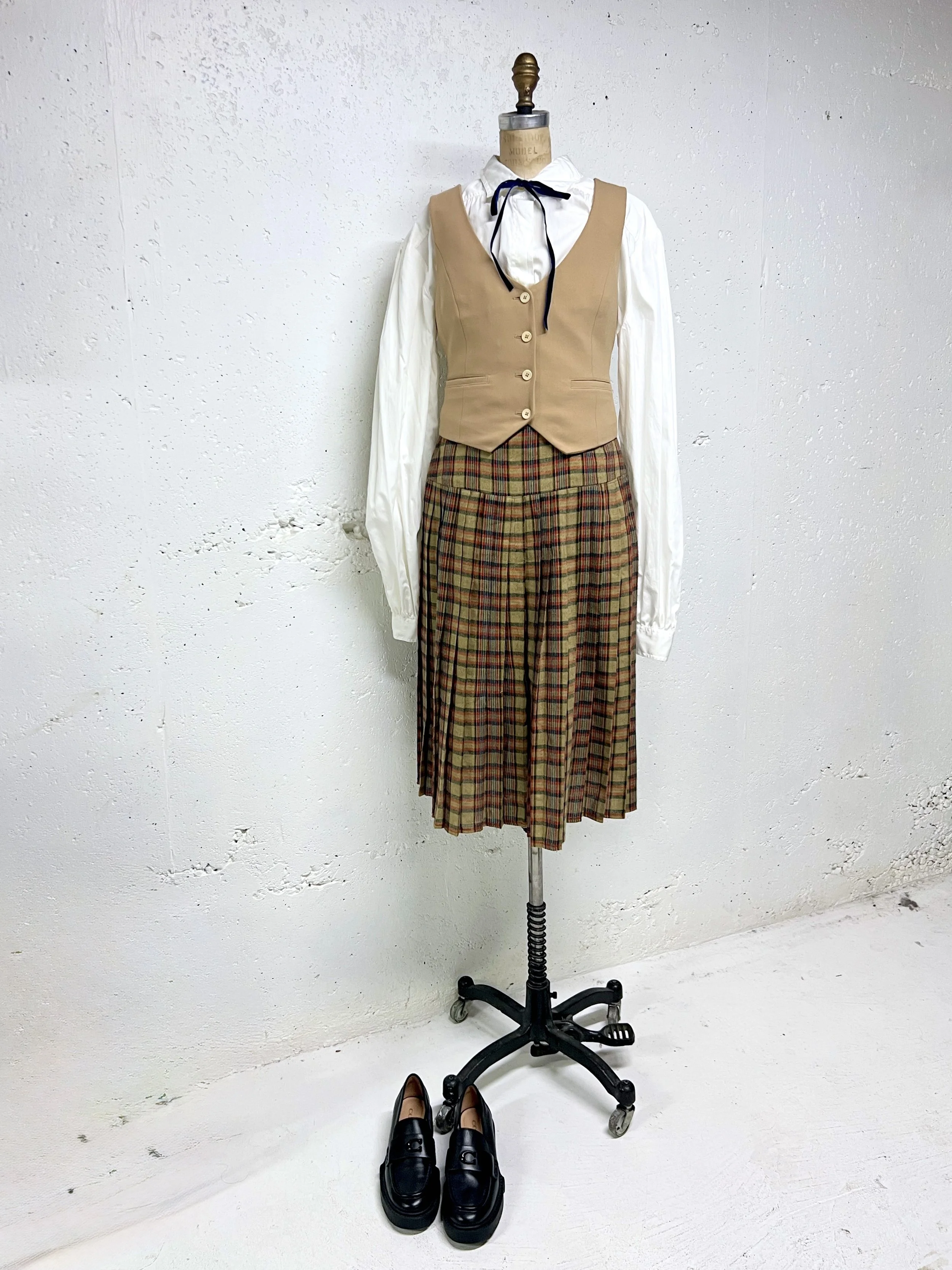 Mannequin wearing vintage-inspired schoolgirl outfit with pleated plaid skirt, beige vest, white blouse with bow tie, and black shoes, against a plain white wall.