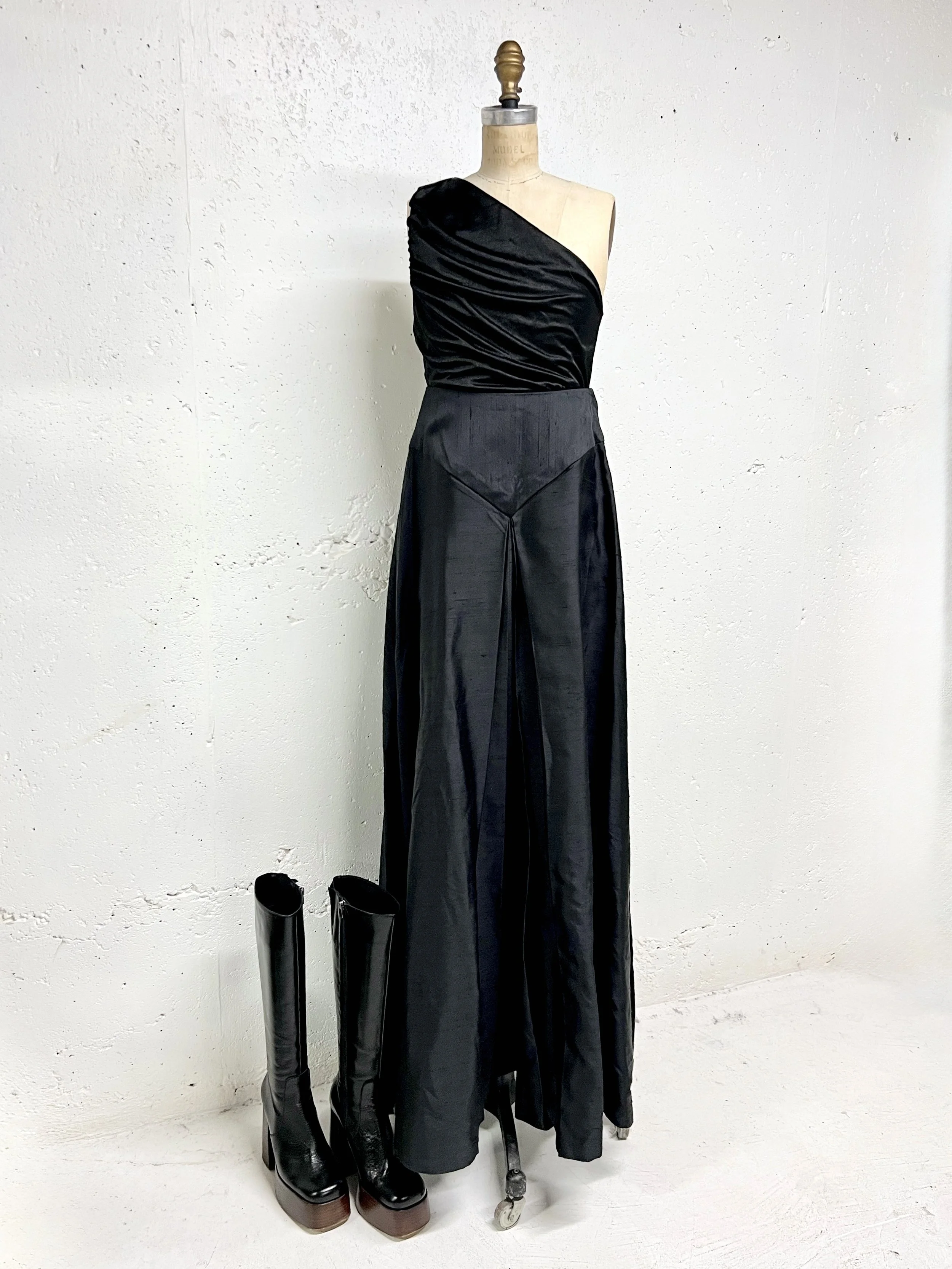 A mannequin dressed in a one-shoulder black satin top paired with high-waisted black satin wide-leg pants, with a pair of tall black platform boots placed next to it.