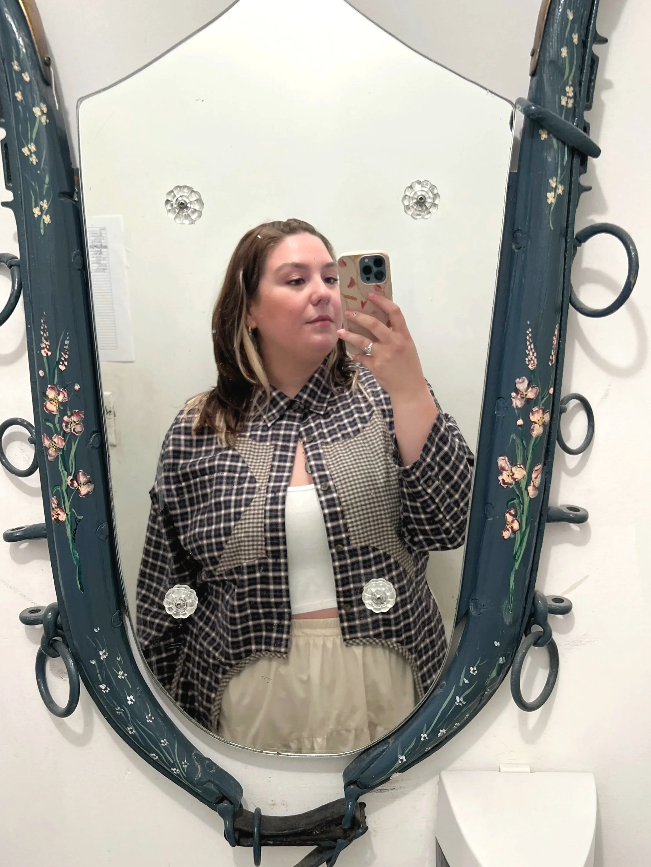 A woman taking a mirror selfie with her phone. She's wearing a checked patterned jacket over a white top, with beige pants. The mirror has a decorative frame with floral designs and hooks.