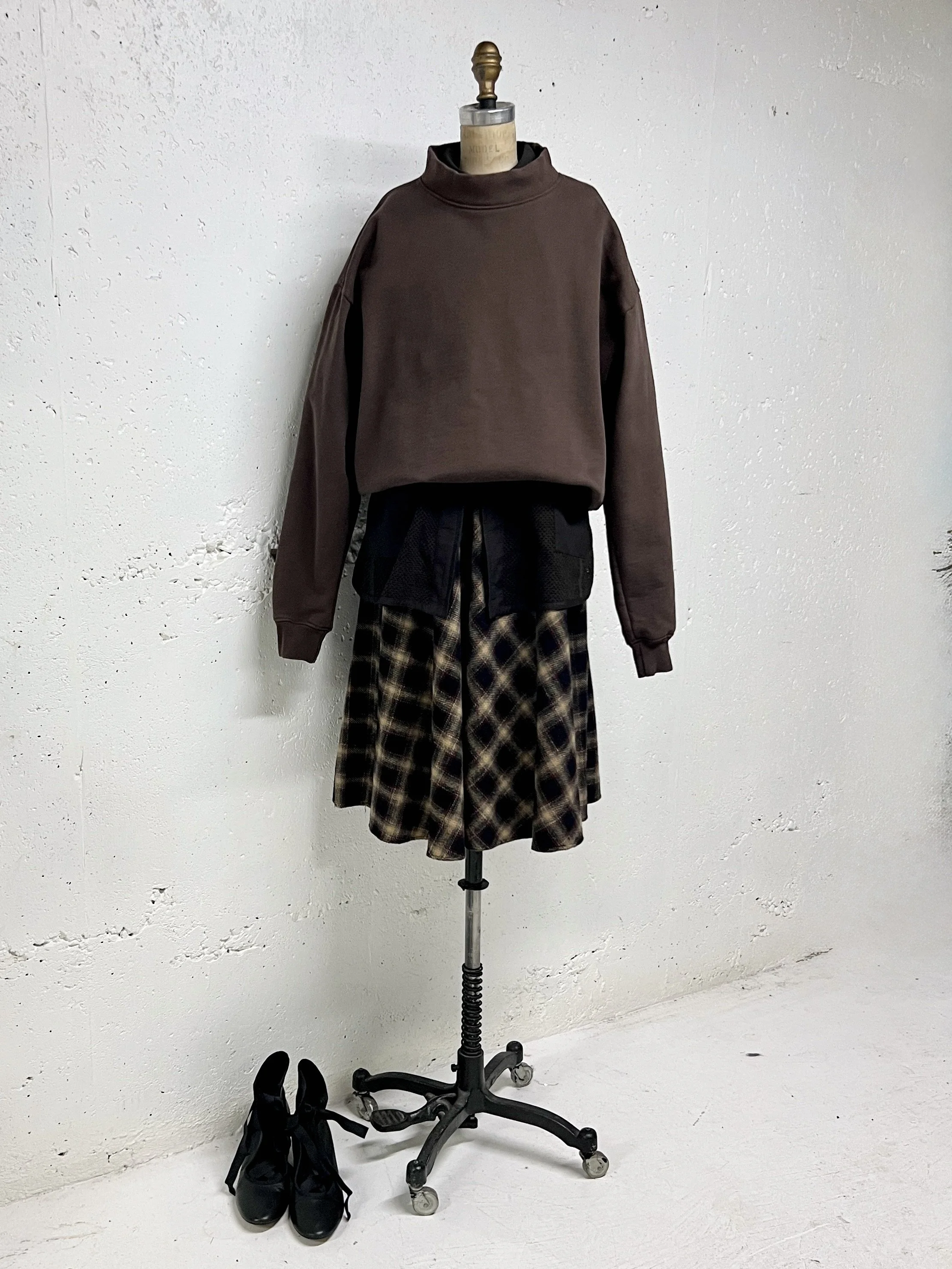 Clothing display featuring a mannequin dressed in a brown oversized sweatshirt, black jacket tied around the waist, and a plaid skirt. Black shoes placed on the ground beside the mannequin.