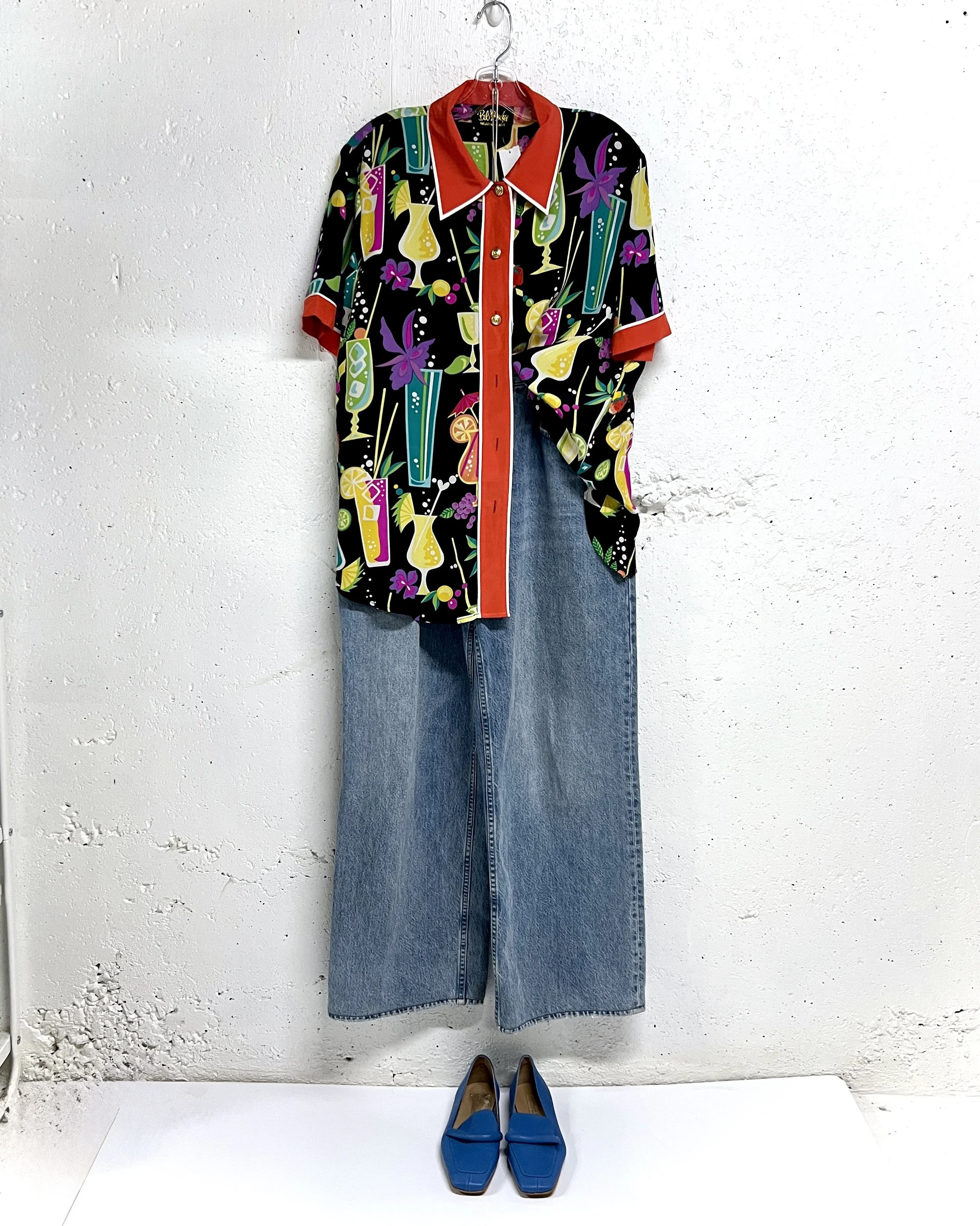 A colorful short-sleeved button-up shirt with a cocktail-themed print, hanging on a hanger against a white wall. Below the shirt are wide-leg blue jeans and bright blue shoes placed on the floor.
